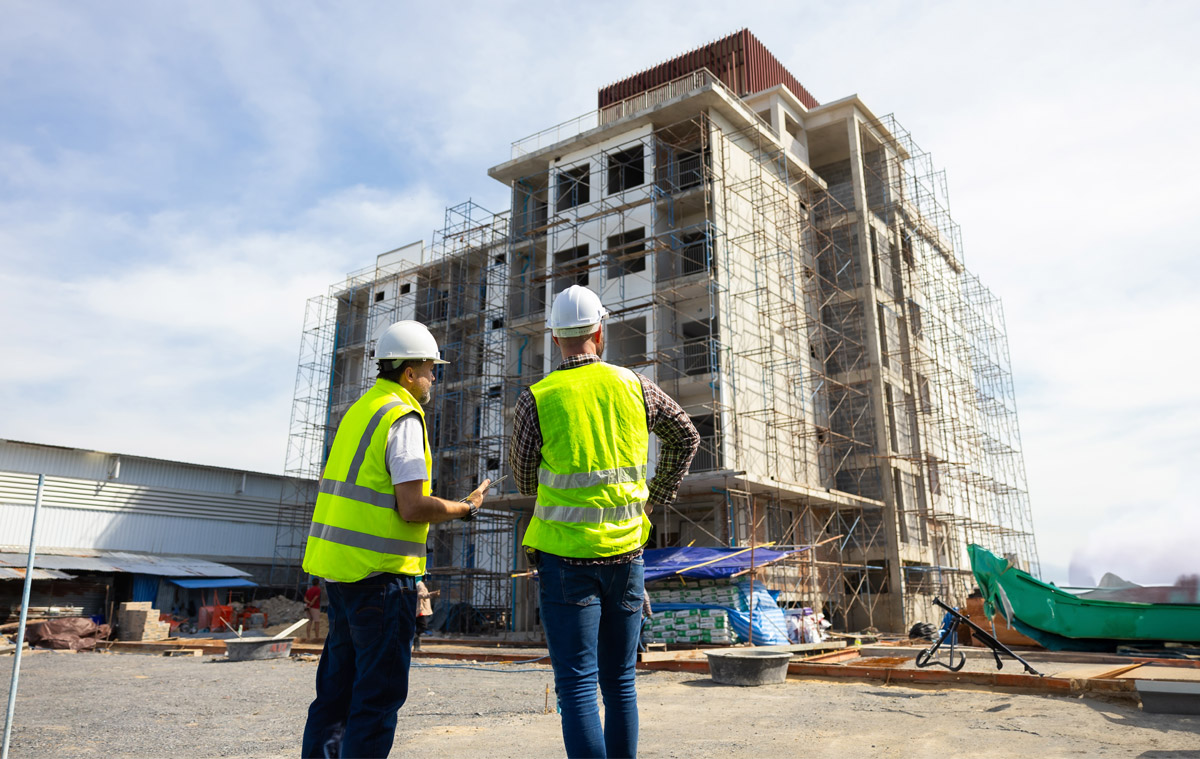 Zwei Bauleiter mit Warnwesten und Helmen begutachten die Fassade eines mehrstöckigen Rohbaus mit Baugerüst auf der Baustelle.