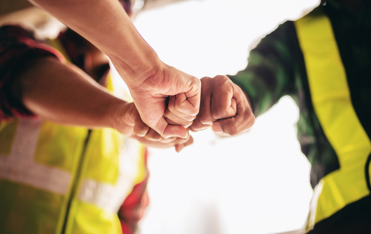 Drei Bauarbeiter mit Warnwesten stoßen zur Teambegrüßung die Fäuste zusammen.